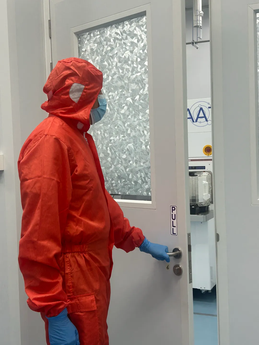 Gowned AAT engineer entering the Yishun cleanroom tool bay, with an AAT atomic layer etch tool beyond the door